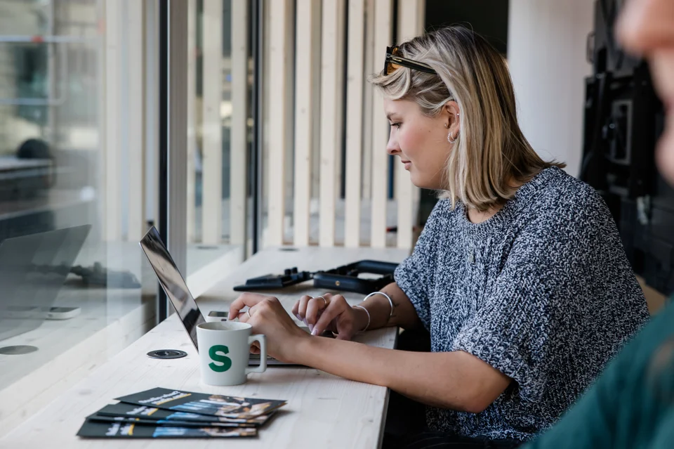 Junge Frau sitzt im studio_one-Open Space und arbeite am Laptop, während sie einen Kaffee trinkt