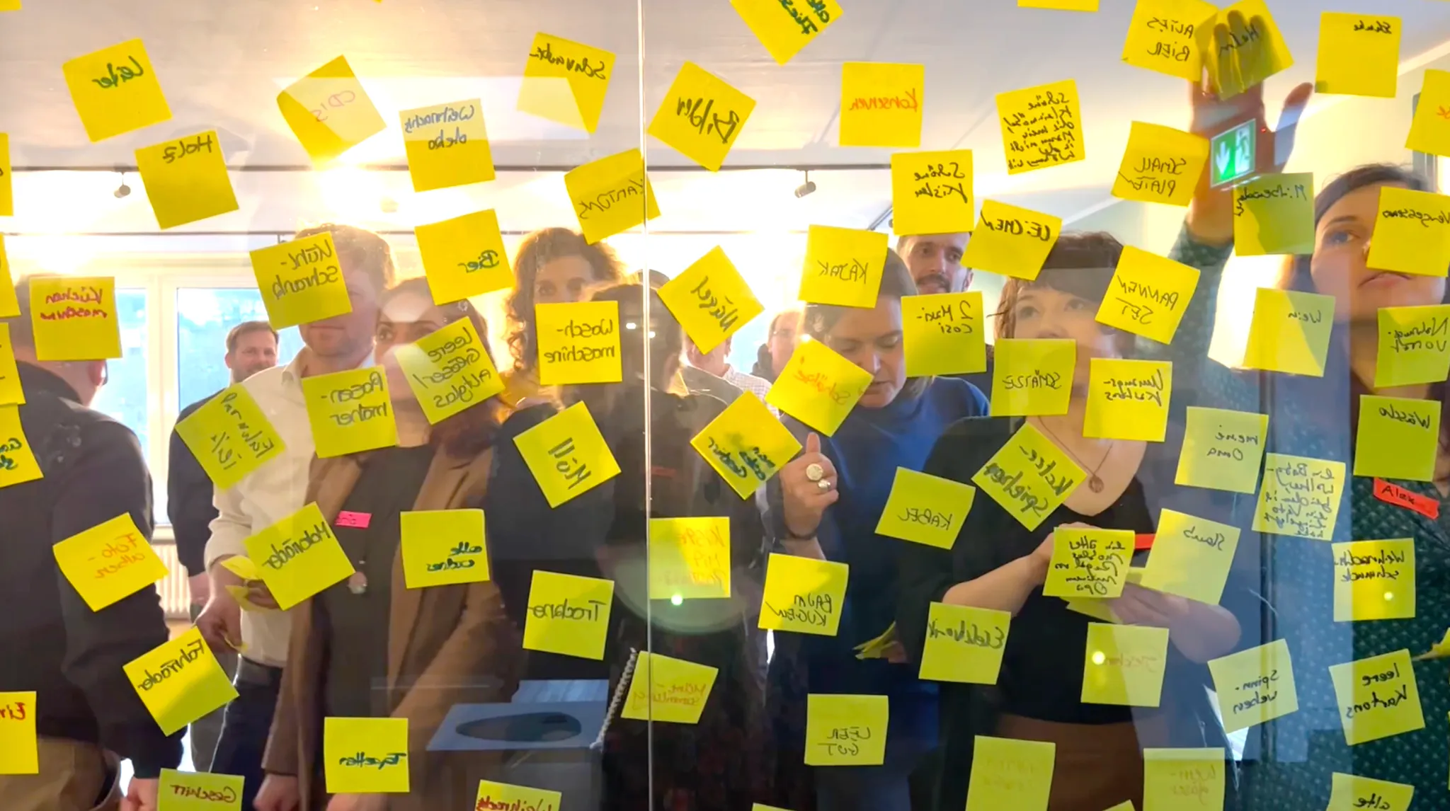 Durch eine Glasscheibe fotografierte Menschen, die im Zuge eines Workshops im studio_one beschriftete Post-It's auf die Scheibe kleben